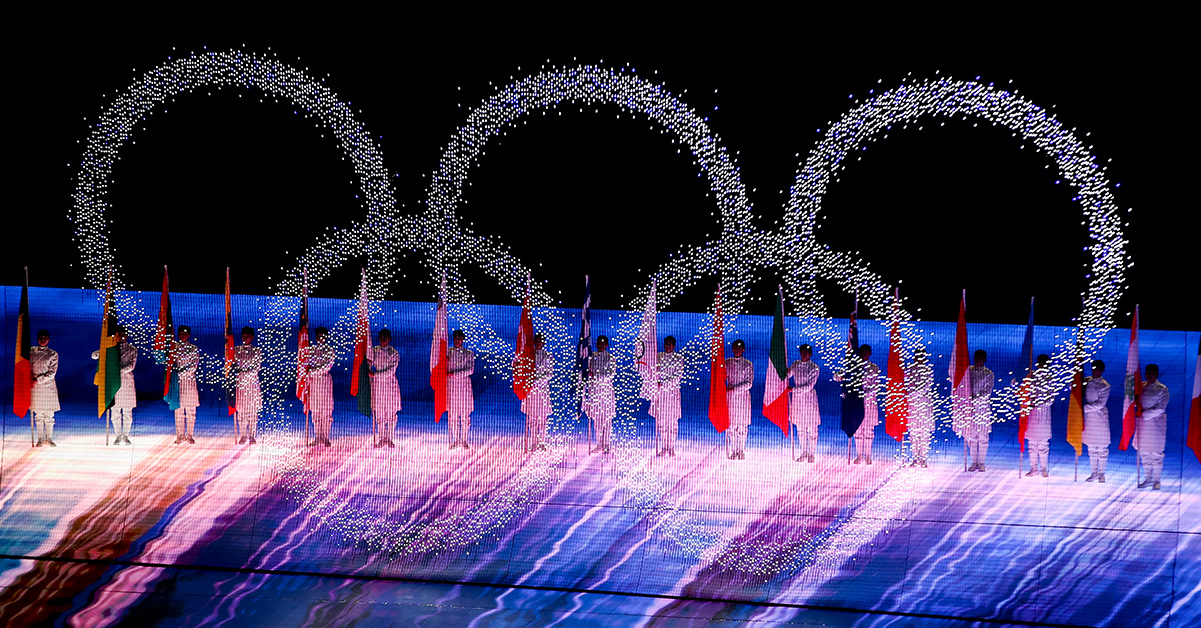 Stor ljusdisplay av de olympiska ringarna. I bakgrunden står människor med nationsflaggor.